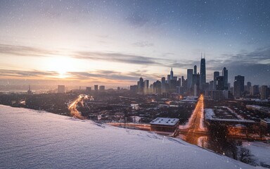 A snowy cityscape at dusk with a highway leading towards the skyscrapers and a bright, warm sunset in the distance.