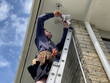 © Harriet - Caucasian man wearing a leather tool belt standing on a ladder installing a security sensor light to the underside of a soffit on the corner of a brick clad house in New Zealand