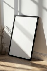 Sunlit minimalist black-framed blank canvas leaning against a white wall with shadows and wooden floor.