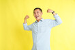© Wayan - Asian man in light blue denim shirt with strong gesture and raising arm showing biceps isolated on yellow background