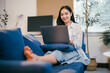 © Parichat - Happy asian woman working from home on her laptop, sitting comfortably on a sofa, embodying a modern lifestyle