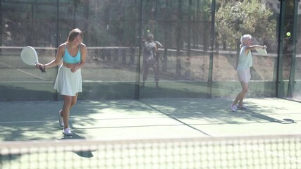 Wall Mural - Senior woman in shorts playing padel tennis on court with young woman. Racket sport training outdoors. 