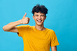 © SHOTPRIME STUDIO - cheerful young man giving a thumbs up with a big smile, wearing a yellow t shirt on a bright blue background, radiating positivity and confidence in a casual setting