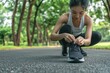 © ChimE - Focused on her legs, an Asian woman ties her shoes in the park, ready for a jog amidst lush greenery.