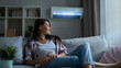 © SS Digital - Woman who's sitting on sofa under warm plaid in living room switches off her air conditioner on wall. Young girl adjusting modern AC system, regulating temperature and enjoying cool