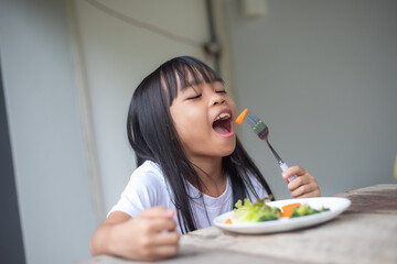 Wall Mural - A young girl is sitting at a table with a plate of vegetables in front of her.