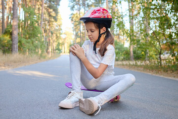 Pretty little girl learning to skate on beautiful summer day in park wearing safety helmet and hurt her knee while riding sitting on asphalt road and touching painful leg