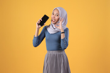 Wall Mural - happy asian indonesian muslim woman holding smart phone and showing smart phone blank screen and giving ok finger gesture on isolated yellow background