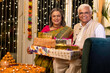 © G-images - Happy senior husband giving surprise gift to excited wife in illuminated home on occasion of Diwali festival