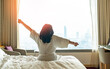 © Chinnapong - Hotel relaxation on lazy day with Asian woman waking up from good sleep on bed in weekend morning resting in comfort bedroom looking toward city view, having happy, work-life quality balance lifestyle