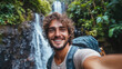 © Mark - Smiling backpacker capturing a selfie with a large waterfall behind him in a lush tropical forest, surrounded by vibrant greenery on an outdoor adventure