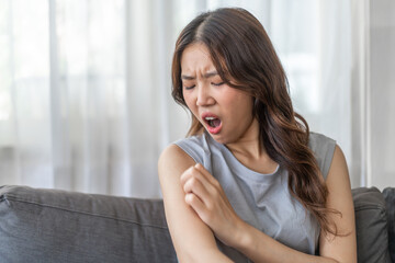 Asian woman with a disgusted expression touching her arm, feeling discomfort or itchiness on a sofa, health and skin irritation issue, unpleasant feeling, young female showing reaction of sensitivity