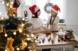 © sofiko14 - Caucasian mother and daughter wearing Santa hats baking together. They knead dough in cozy kitchen filled with holiday decorations.