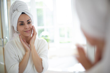 Wall Mural - Woman with a towel wrapped around her head, wearing bathrobe, smiles as she touches her face in front of bathroom mirror. Bright natural light fills the room, creating a serene, spa-like atmosphere