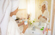 © My Ocean studio - Woman in bathrobe and towel on her head smiles in front of bathroom mirror, holding a jar of skincare cream. The counter features various beauty products, creating a relaxed and fresh morning vibe