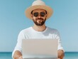 © MNFTs - A happy man wearing a straw hat and sunglasses, working on a laptop on the beach under a clear blue sky.