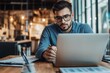 © fotofabrika - Young man with glasses focused on his laptop in a modern workspace during the daytime, surrounded by notebooks and stationery