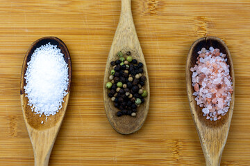 Three wooden spoons arranged on a bamboo cutting board.  Spoons filled with pretzel salt, whole peppercorns, and himalayan salt.