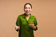 © Maria Vitkovska - Positive, happy Asian woman wearing green shirt smiling, blogger recording video on beige background