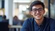 © Maria Mikhaylichenko - Confident young professional with a warm smile, wearing glasses and a blue sweater, in a modern office environment. Blurred background suggests a busy workspace.