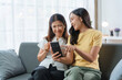 © amnaj - Young woman is showing something funny on her smartphone to her mother while sitting on a sofa at home