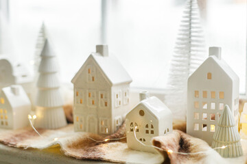 Wall Mural - Christmas little ceramic houses, snowy trees and lights on cozy scarf on window. Xmas background. Merry Christmas! Christmas scene, miniature holiday village. Holiday modern decorations