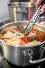 close-up of a hand using tongs to cook soup in stainless steel pot on a stove. Steam rises from the boiling soup, creating a warm and inviting kitchen scene. Perfect for culinary and cooking themes.