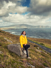 Wall Mural - A teenager girl in a yellow jacket stands on a hill overlooking the ocean and stunning Irish nature scenery. The view is serene and peaceful. Achill island, Ireland. Travel and tourism.