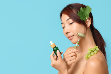 Wall Mural - Beautiful young Asian woman with bottle of cosmetic grape seed oil and face massage roller on blue background