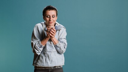 Wall Mural - Happy woman sipping coffee from a cup against blue background, enjoying a hot caffeine refreshment in studio. Confident cool person with modern outfit drinking a beverage. Camera B.