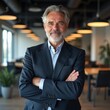 © Makaron - A confident businessman with a gray beard, standing in a modern office setting. Ideal for corporate, leadership, or professional development themes.