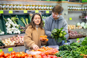 Couple of young woman and young guy choose carrots and spinach in vegetable shop