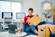 © Parichat - Young asian woman is sitting on her couch and writing on a cardboard box, preparing it for shipping. She is working from home