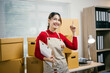 © Parichat - Female business owner is holding a clipboard and pointing up with her index finger while surrounded by cardboard boxes