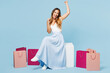 © ViDi Studio - Full body young happy excited woman wear casual clothes sits near paper package bags after shopping do winner gesture clench fist isolated on plain blue background. Black Friday sale buy day concept.