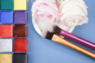 brushes on coaster with face painting palette and artificial flower on blue background