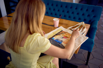 a beautiful young artist in a yellow dress paints a customized shopping bag