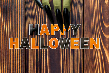 Wall Mural - Green witch hands with long black nails showing black and orange letters saying Happy Halloween on a wooden background