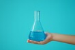 © RaptorWoman - Hand holding a laboratory glass flask filled with blue liquid against a vibrant blue background, symbolizing science and research.