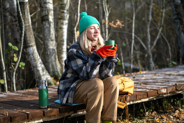 Satisfied middle aged woman spending tranquility time alone walking in autumn forest dreaming, drinking hot thermos tea. Relaxed calm female rest on wooden path enjoys late fall park, stress free