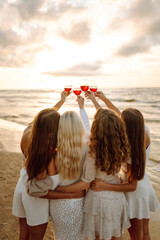 A group of four women friends, dressed in summer outfits, stands on the beach at sunset. They raise their glasses in a toast, celebrating their friendship by the ocean. Hen party on the beach. 