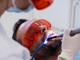 A patient is lying in a dental chair while a dentist works on their teeth using a laser tool for teeth whitening. Protective eyewear shields the patient's eyes from the bright light during the procedu