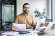 © Liubomir - Male entrepreneur with glasses reviewing papers at desk in bright office. Smiling person analyzing reports with laptop and phone on table. Business setting includes calculator and open documents.
