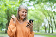 © Liubomir - Mature woman expressing happiness while using smartphone outdoors. Wearing orange sweater, celebrating news, showing joy in park setting. Senior lady enjoying moment, surrounded by lush greenery.