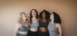 © DBFilms - Group of four women of different body shapes, standing together, laughing and bonding in workout outfits.