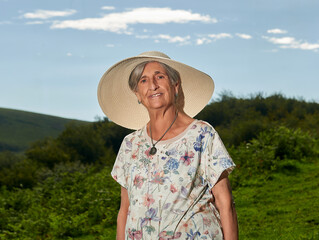 An octogenarian woman with a sun hat poses in front of a meadow with a mountain in the background and a clear sky