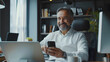 © sakir - Happy mature 45 years old business man executive sitting at desk using mobile phone. Smiling middle aged businessman looking at camera working on computer technology holding smartphone. generative ai.