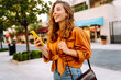 © maxbelchenko - Cheerful young woman with curly hair enjoys a sunny day while walking in an outdoor city area and using her smartphone. Lifestyle, travel, tourism, technology, blogging, communication concept.
