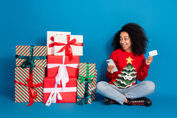 Poster - Full body photo of young woman look pile gifts hold device credit card dressed red christmas clothes theme party on blue color background