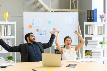 Young Asian businesswoman and businesswoman sitting working at office desk with laptop celebrating goal achievement raise hand up in the air. Businesspeople successful and enjoy work.